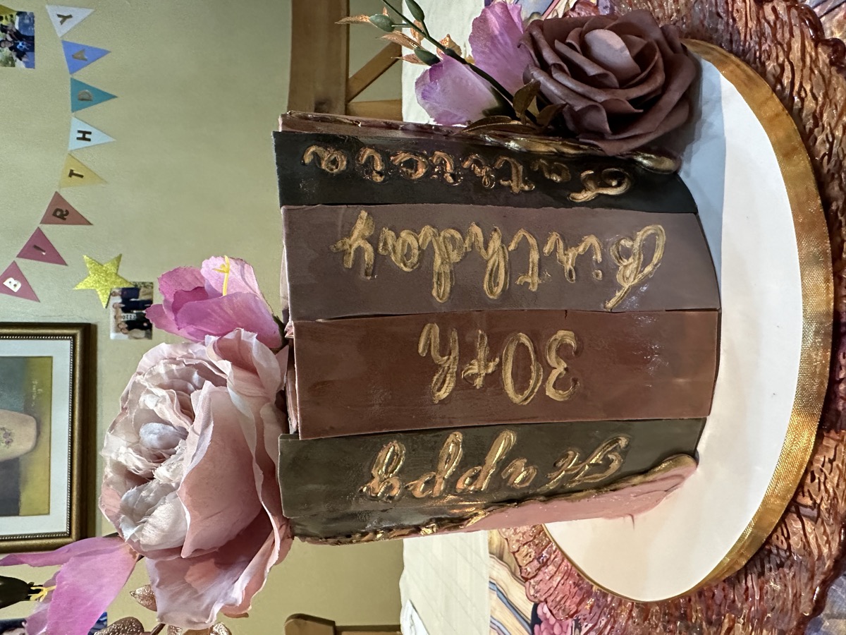 Chocolate book-shaped birthday cake with gold lettering and silk flowers